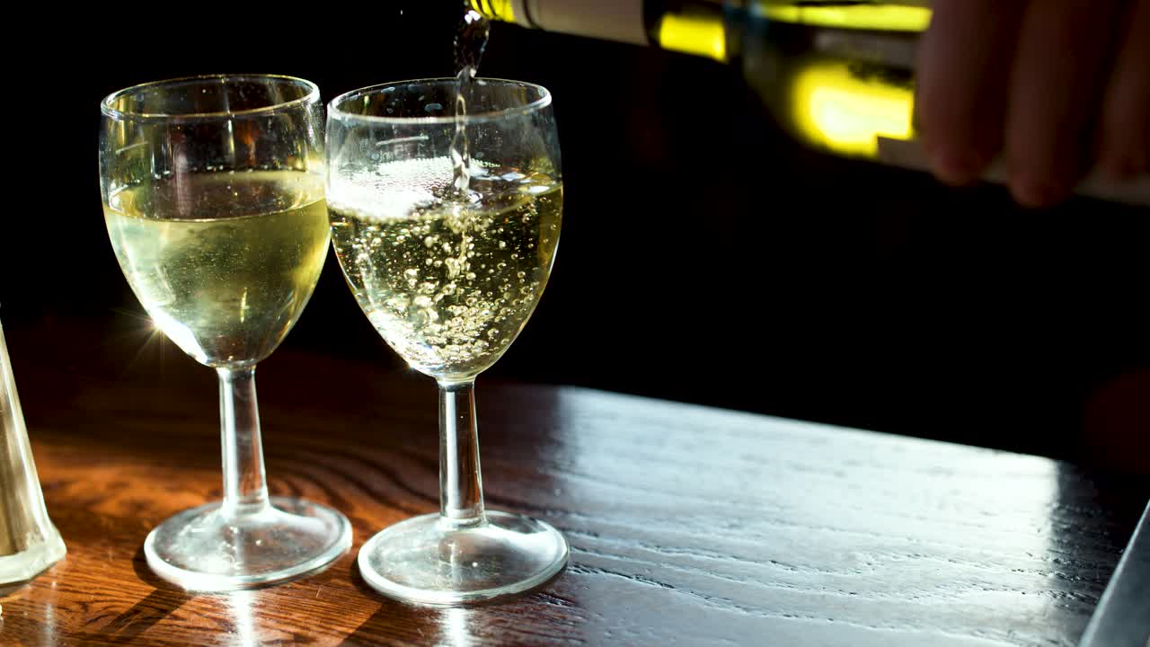 Hand pours sparkling white wine into glassware on wooden table, warm lighting, close-up view