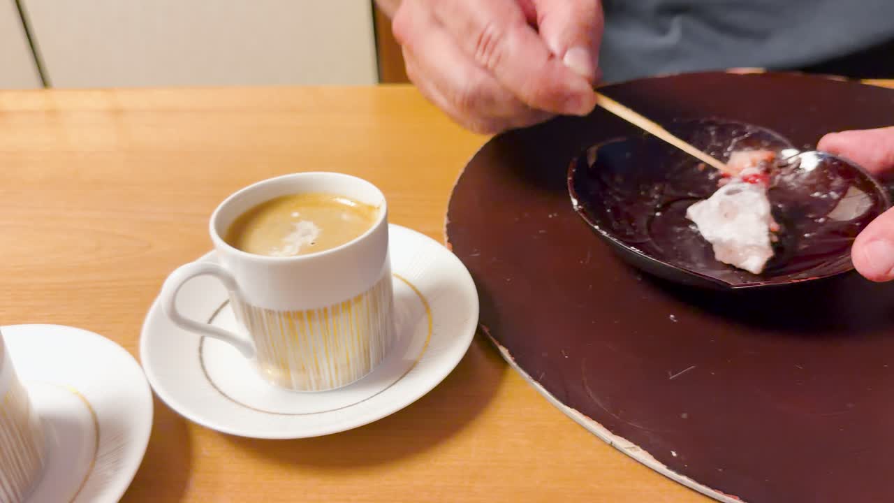Gros plan de mains âgées utilisant un pic en bois pour manger du mochi à la fraise à côté d'un espresso.