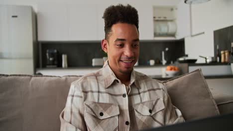 A-happy-young-Black-skinned-brunette-man-with-stubble-in-a-checkered-beige-shirt-sits-on-a-modern-gray-sofa-and-works-on-his-laptop-remotely-while-at-home-in-a-studio-apartment