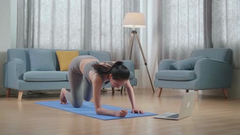 joven atleta asiática en ropa deportiva haciendo yoga en el antebrazo en la postura de tabla en la alfombra en casa con el entrenador en línea a través de una computadora portátil