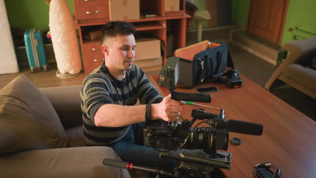 Studio guy adjusting camera lens on tripod in home studio while preparing for video shoot. Camera gear and microphone arranged on table, ready for filming project