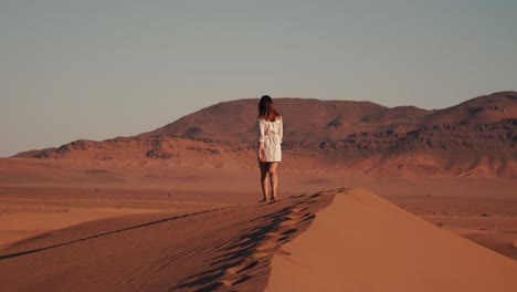 Young-caucasian-woman-walking-on-top-of-a-desert-dune-during-sunset-with-mountains-in-the-background-in-Zagora,-Morocco