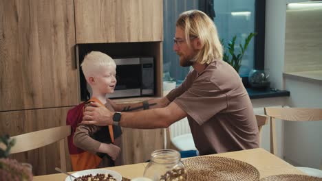 un hombre rubio con gafas y barba abraza a su pequeño niño albino de cabello blanco que se puso una mochila roja antes de su día en la escuela. niño feliz se despide de su padre y va a la escuela