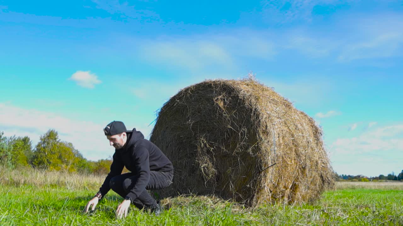 Man with black clothing jumps off from round hay bale in slow motion making the hay move and fall in slow motion during summer time while sun is shining in a farm field that has blue sky in the back.