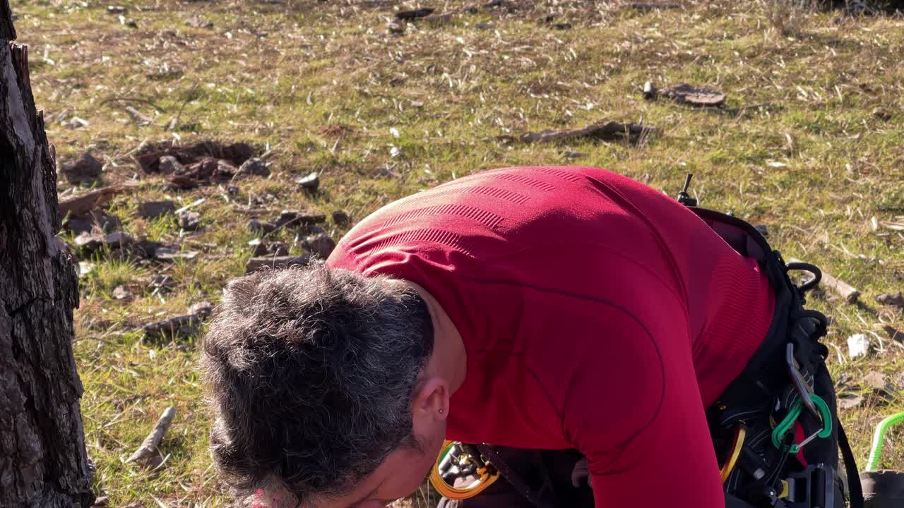Starker Mann in roter Kleidung mit Klettergurt schärft Kettensäge im Wald.