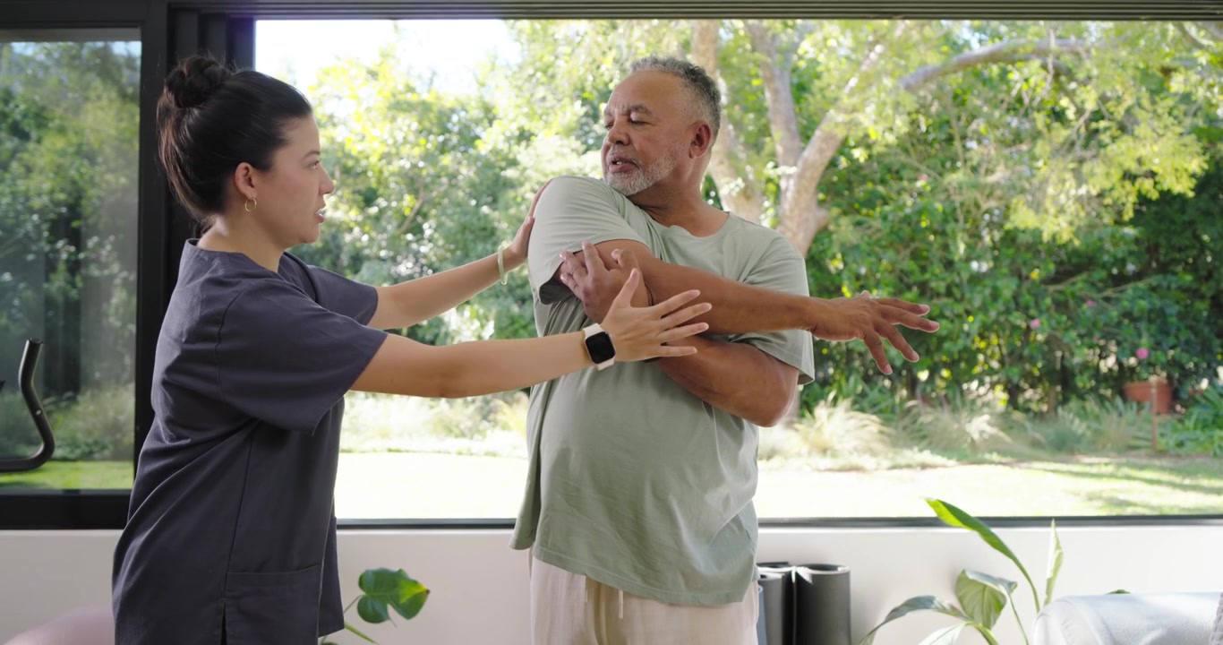 Senior African American man following Asian therapist reaching guiding shoulder for rehab by window