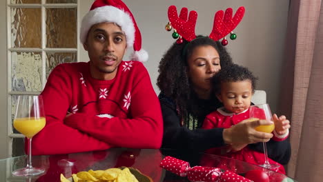 Young-Parents-with-Small-Child-On-a-Christmas-Video-Call-Listening-and-Talking-to-Camera