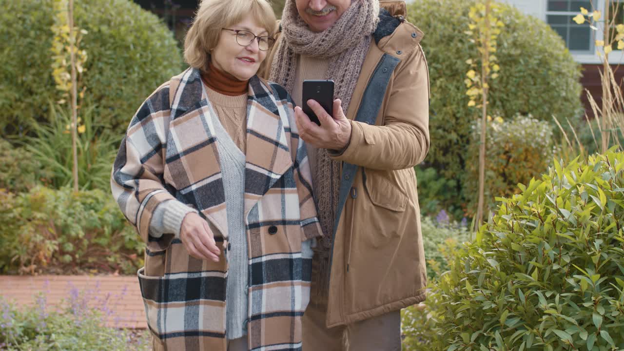 정원과 집에 대한 화상 통화에서 스마트폰으로 인사하는 세련된 따뜻한 캐주얼웨어를 입은 명랑한 노인