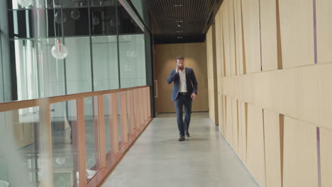 an attractive young, bearded businessman in a blue suit has a business conversation as he enters the offices through a modern wooden hallway 1