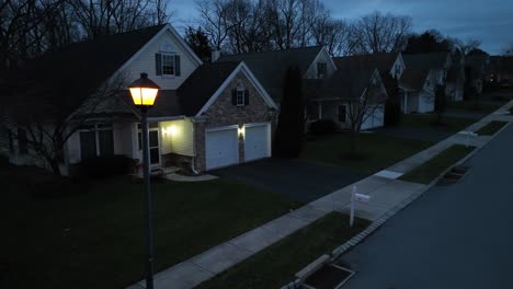 twilight, suburban street lined with detached homes, lamplight glowing warmly