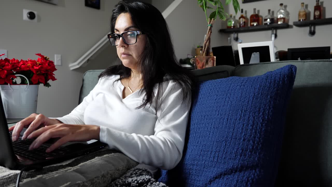 mujer escribiendo en la computadora portátil mientras se sienta en el sofá en la sala de estar, enfocada en el trabajo