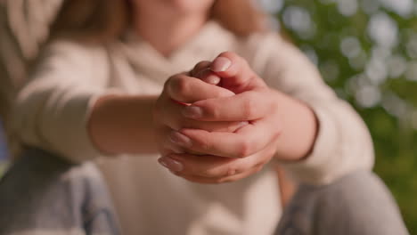 young girl sits outdoors, hands clasped together in quiet contemplation, with blurred background featuring greenery and sunlight casting a soft glow