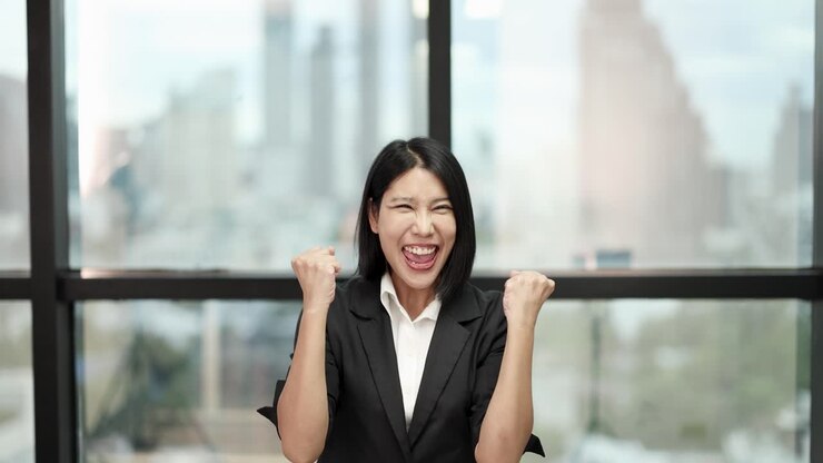 Asian businesswoman in suit celebrates achievement, smiling and raising fists in bright office environment