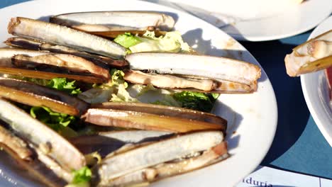 Weibliche-Hand-Nimmt-Gegrillte-Meeresmuscheln-Auf-Einem-Weißen-Teller-Mit-Salat