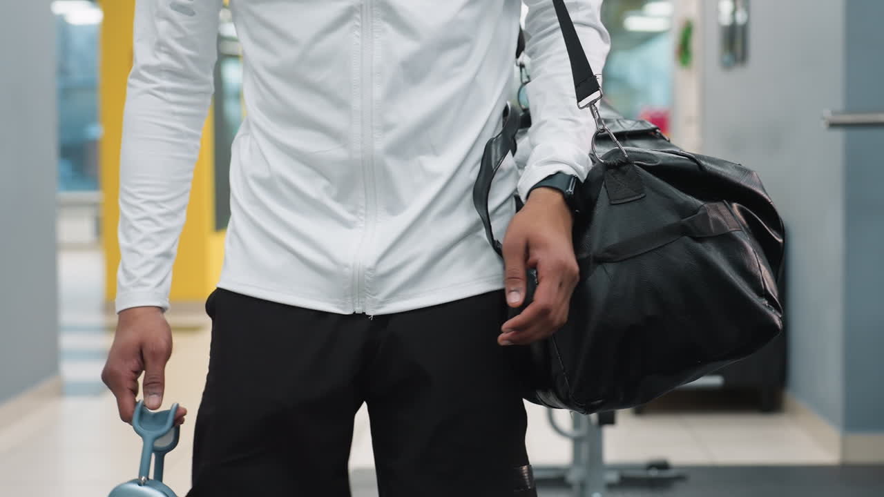 close up of athlete carrying black duffel bag and wireless headphones walking through bright modern gym past weight bench and equipment reflecting in glass as natural light filters through windows