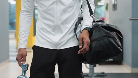 close up of athlete carrying black duffel bag and wireless headphones walking through bright modern gym past weight bench and equipment reflecting in glass as natural light filters through windows