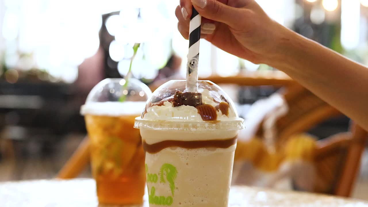 A hand stirs a creamy iced drink with caramel topping in a sunlit Gold Coast cafe setting