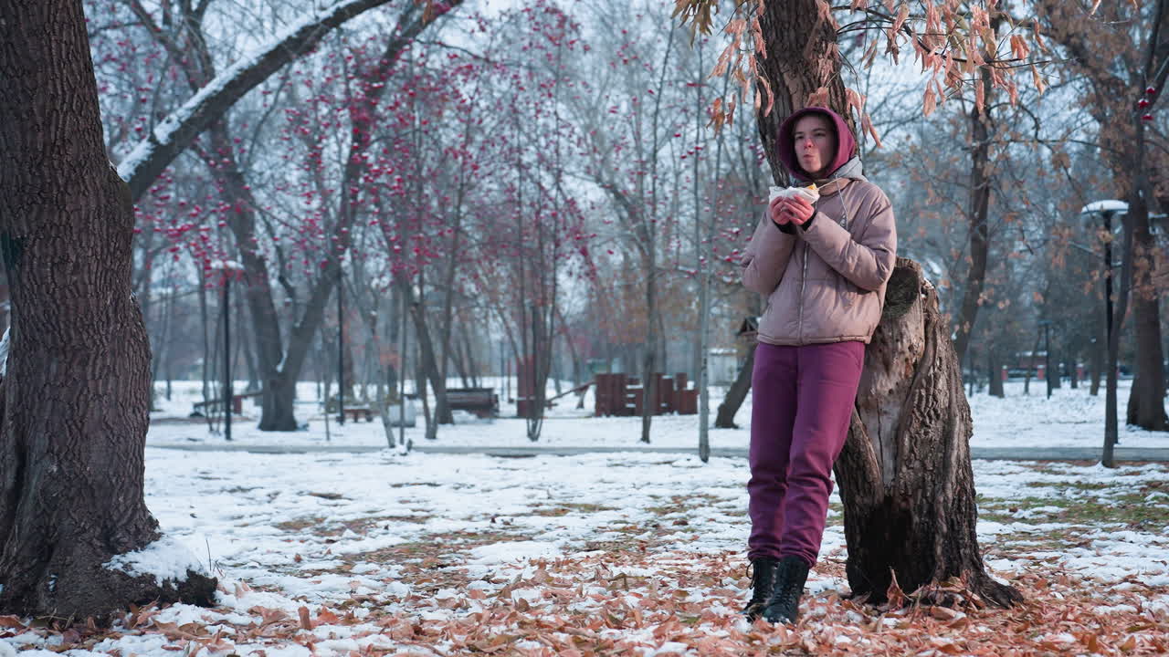 dama con traje de invierno descansando en un árbol comiendo algo en un parque al aire libre durante el invierno, reflejo de luz del fondo, disfrutando de la comida, entorno de invierno, estado de ánimo relajado, tiempo de ocio en el parque, clima frío
