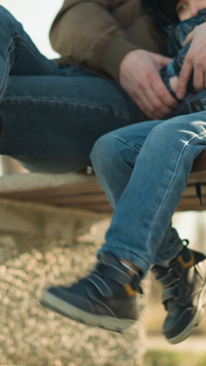 벤치에 가까이 앉아있는 어른과 아이의 클로즈업, 아이는 놀이로 다리를 흔들고, 어른의 다리를 부드럽게 때리며, 나무와 전기 기둥의 흐릿한 배경