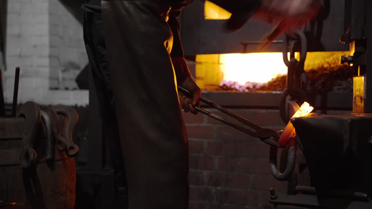 Blacksmith holding a heated chain link with tongs while shaping it in a forge workshop without visible sparks