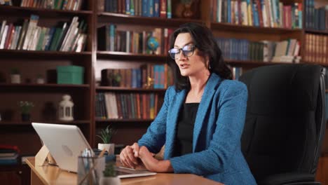 la mujer está sentada en el interior con una computadora portátil en la mesa, rodeada de muchos libros