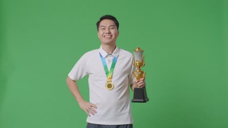 asian man with a gold medal and trophy smiling to camera with one arm akimbo posing as the first winner on green screen background in the studio