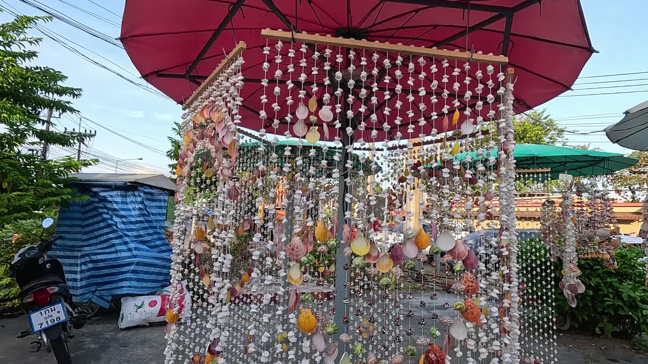 Vibrant shell decorations sway under a red umbrella in a bustling Phuket market, captured with smooth camera movement and natural lighting