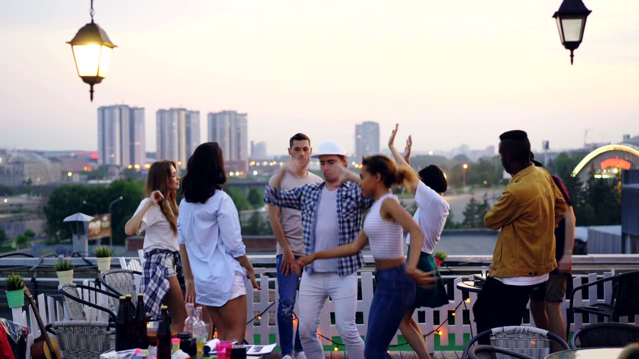 Multiracial group of friends is dancing on rooftop having cool party with DJ in the evening in summer. Joyful youth, entertainment and big city concept.