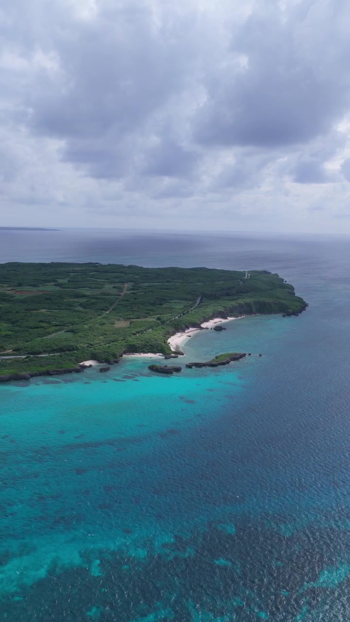 Vista aerea a rotazione lenta del lato più lontano dell'isola di Ikema con spiaggia, acqua blu, cielo nuvoloso e il ponte di Ikema in lontananza, Okinawa, Giappone.