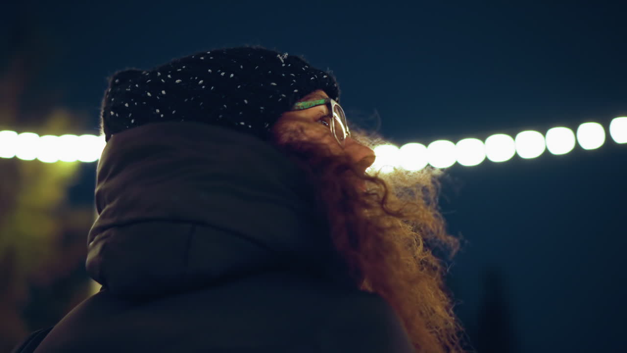 Close up of woman with glasses and curly hair wearing black winter hat looking upward under bright festive lights at night, with visible breath in cold air creating atmospheric winter urban scene