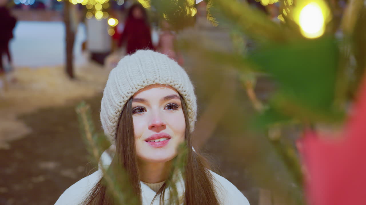 Smiling woman in white beanie and winter coat admiring pine tree with warm holiday glow, soft festive lights highlight her face while people pass by in blurred background