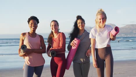 portrait of group of diverse female friends holding yoga mats at the beach