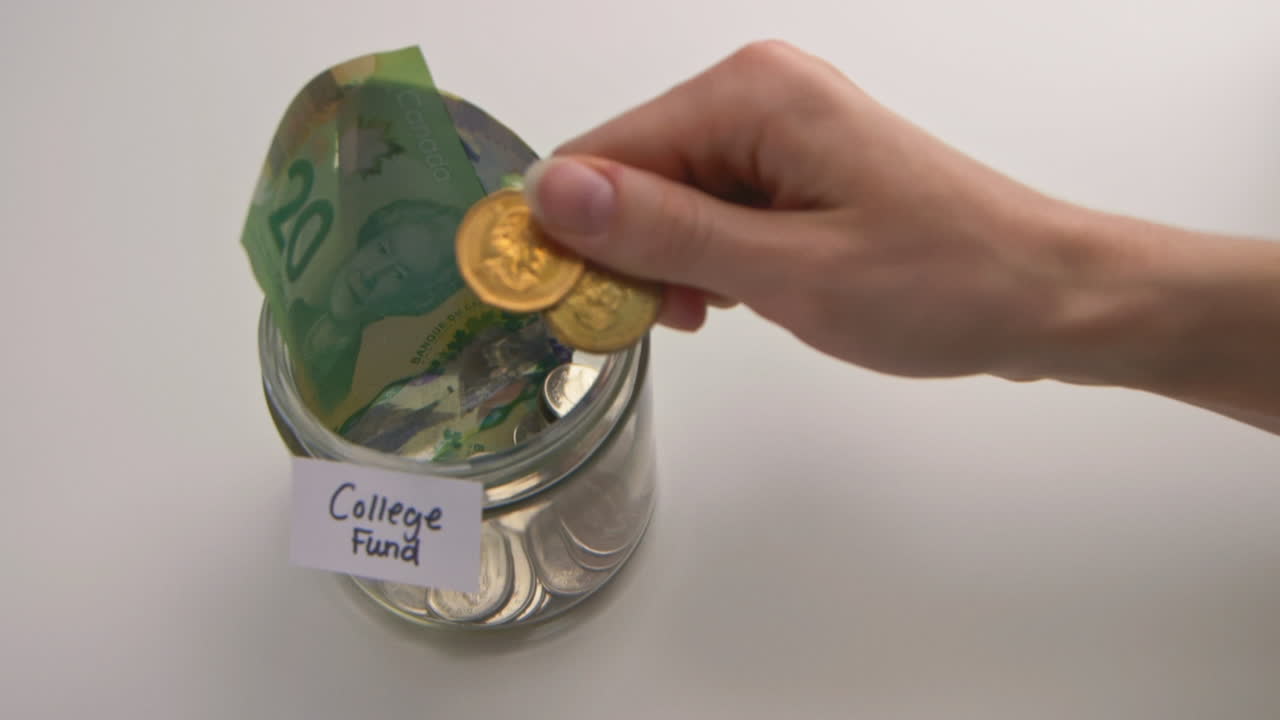 A caucasian woman's hand puts two loonies into a &amp;quot;College Fund&amp;quot; jar