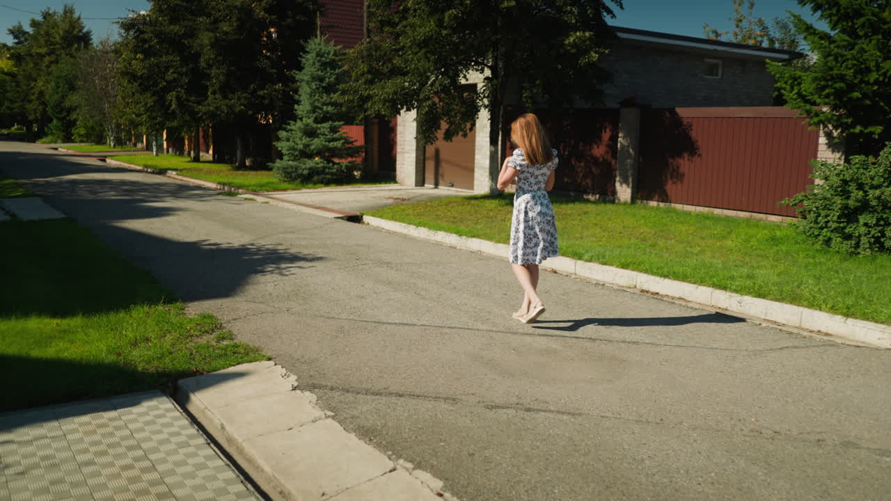Rear view of woman adjusting dress while strolling down peaceful suburban street with green lawns, trees, and residential buildings under bright sunlight during calm sunny day