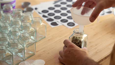 filling a square glass jar with dried chives using a funnel on a wooden countertop
