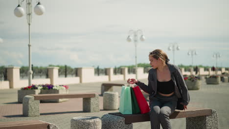 lady in grey clothing drops her shopping bag on an outdoor bench, adjusts a red shopping bag while holding a black handbag sits on the bench, looks to the right with a focused expression