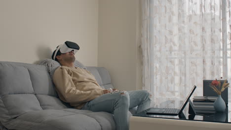 relaxed man enjoying virtual reality at home on couch with controller and laptop nearby