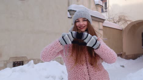 young woman wearing a pink coat and a gray hat makes a heart shape with her hands in the snow