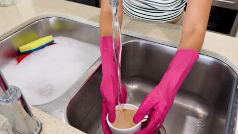 person wearing pink rubber gloves cleans a dirty soap dish under running water in a modern kitchen, using bright lighting and steady overhead camera