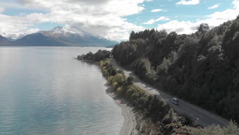 slowmo - campervan driving along the coast of lake wakatipu, queenstown, new zealand with mountains fresh snow in background - aerial drone