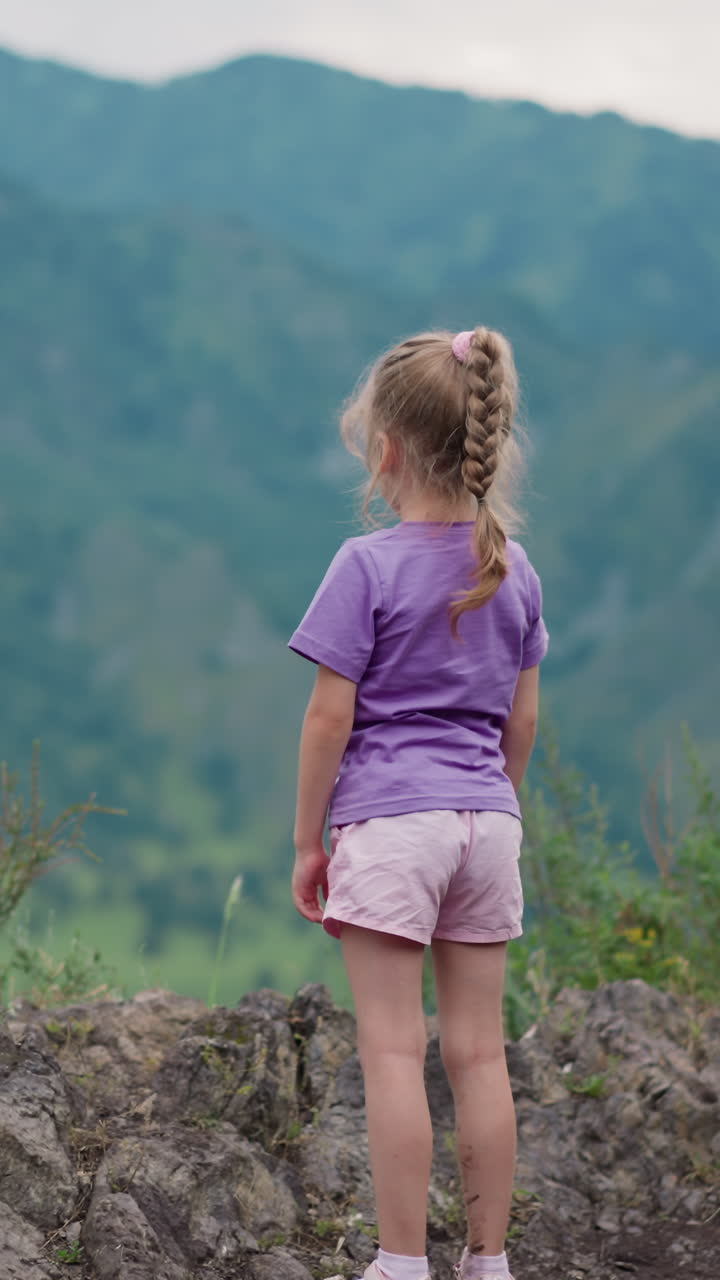 보라색 티셔츠를 입은 포니테일 트레이트를 입은 작은 소녀는 큰 먼 산과 잔디로 인 계곡의 전망을 즐기고 불쾌한 날 뒷면을 느린 동작으로 볼 수 있습니다.