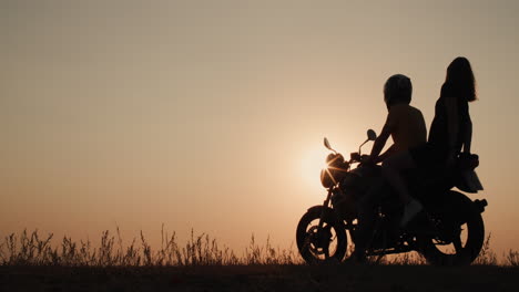 a young couple sits on a motorcycle together admiring the sunset