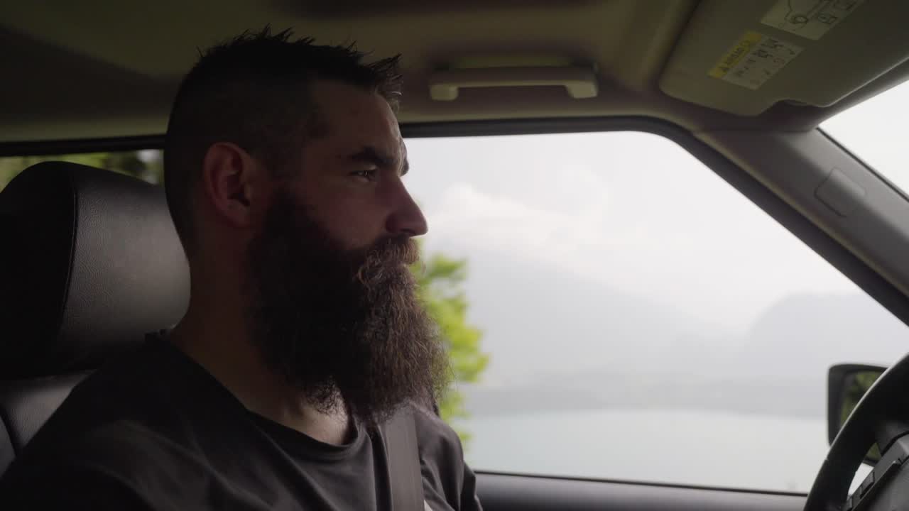 Bearded man driving a car along a mountain road with scenic views outside. The natural light and calm mood suggest a road trip or peaceful travel moment in nature