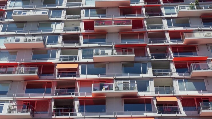 Plano geral estático da fachada de um edifício de apartamentos moderno de vários andares, com inúmeras varandas adornadas com toldos vermelho e laranja brilhantes sob um céu azul claro. A luz solar intensa ilumina o edifício residencial