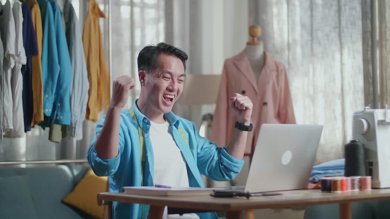 행복한 아시아 남성 디자이너가 노트북으로 작업하고 스튜디오에서 옷을 디자인하는 동안 축하하고 춤을 추고 있습니다.