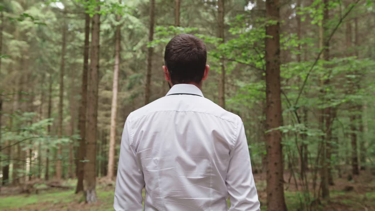 Following shot from behind a man in a white dress shirt as he walks steadily into a dense green forest. The shot evokes a sense of purpose, a journey, or a contemplative escape into nature.