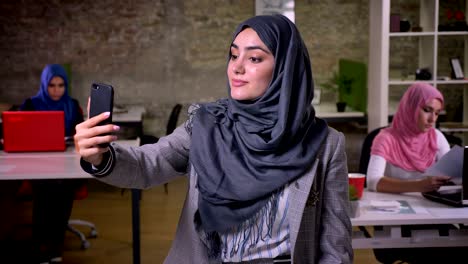 amazing arab female is talking over phone, having wvideo call while standing in brick studio with working middle east girls sitting behind her