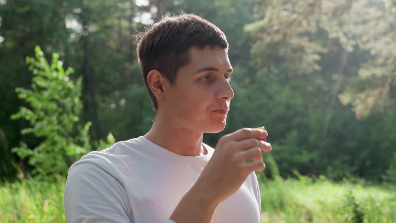 Close up of young man drinking water from cover while standing outdoors surrounded by sunlight, refreshing himself in calm natural environment, showing relaxation, and peaceful summer mood