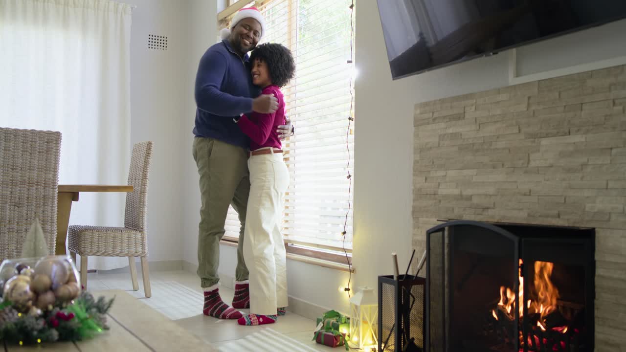 Diverse couple entering cozy living room, gazing out window, dancing and sharing joy near fireplace
