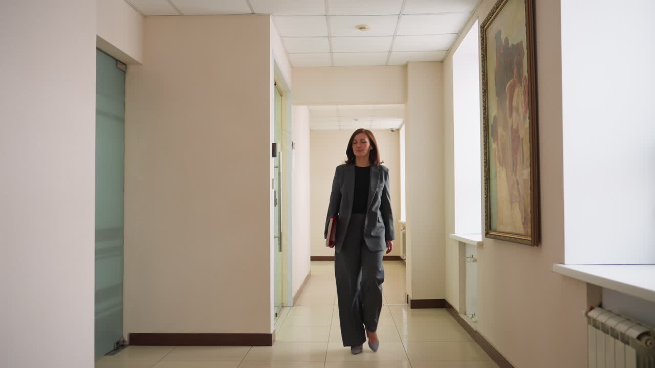 Confident businesswoman walking down bright office hallway holding folder in hand wearing professional dark suit with determined expression surrounded by modern decor